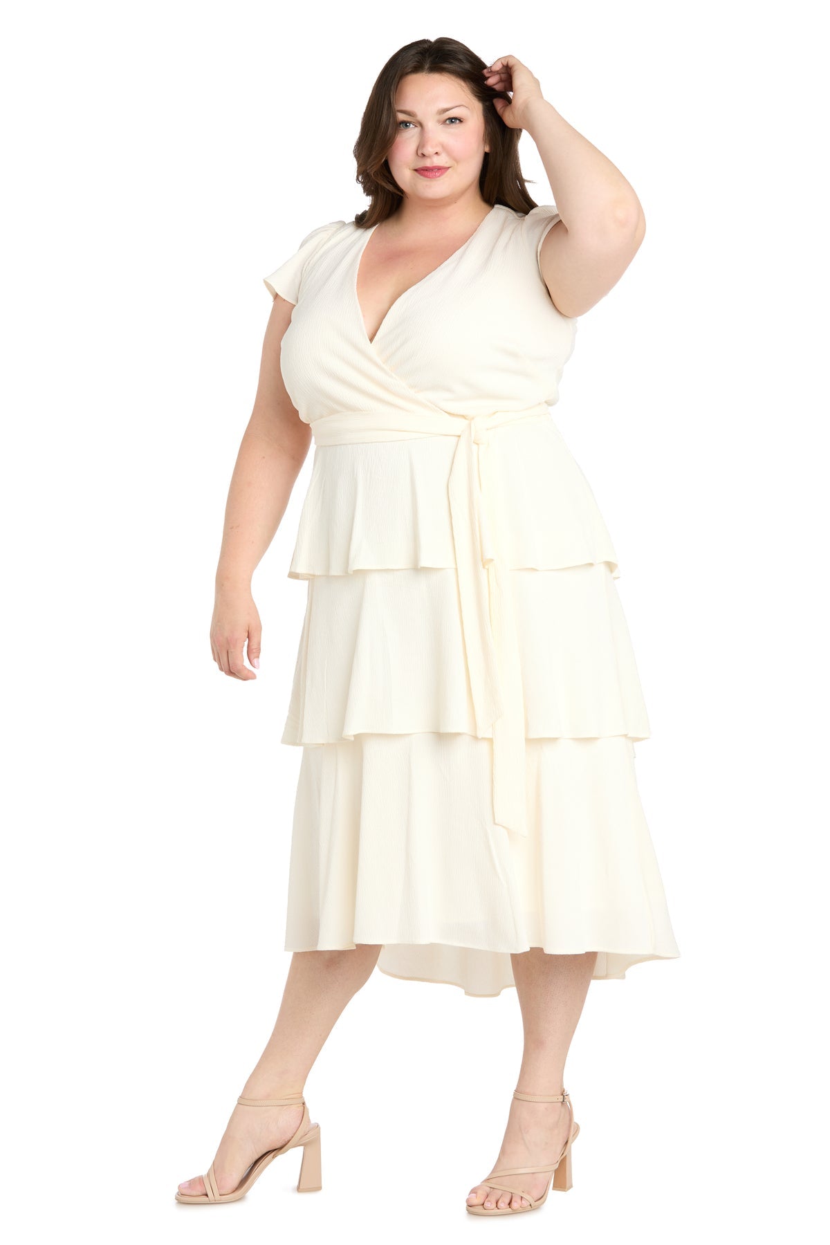 A woman models the Plus-Size White Party Dress: R&M Richards 6677W, featuring short sleeves, a tiered wrap front, and tie waist, styled with nude heeled sandals and her hand in her hair.